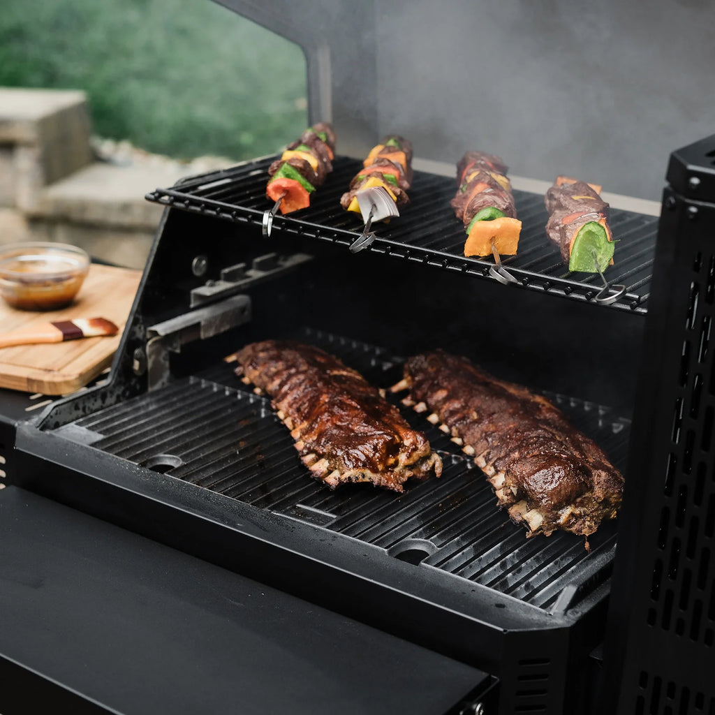 An open grill showing 4 kebab skewers cooking on the warming rack above the  main cooking grate. 2 full racks of ribs cook on the main cooking grate. The grill front shelf is up in front of the main cooking grate. A wooden board with a bowl of sauce and a basting brush on it sits on the side shelf to the left of the grill.