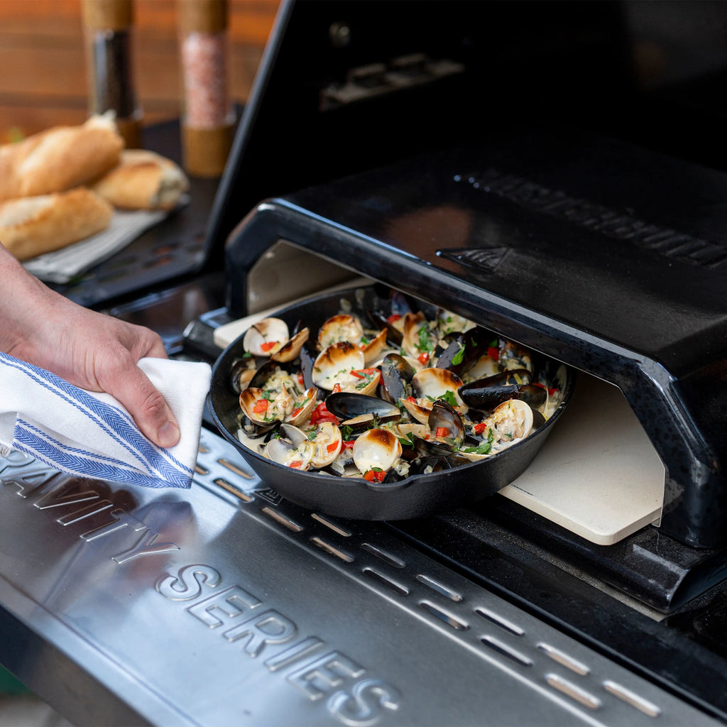 Someone removes a case iron skillet of cooked mussels from the Gravity Series Pizza Oven installed in a Gravity Series 1150 grill.