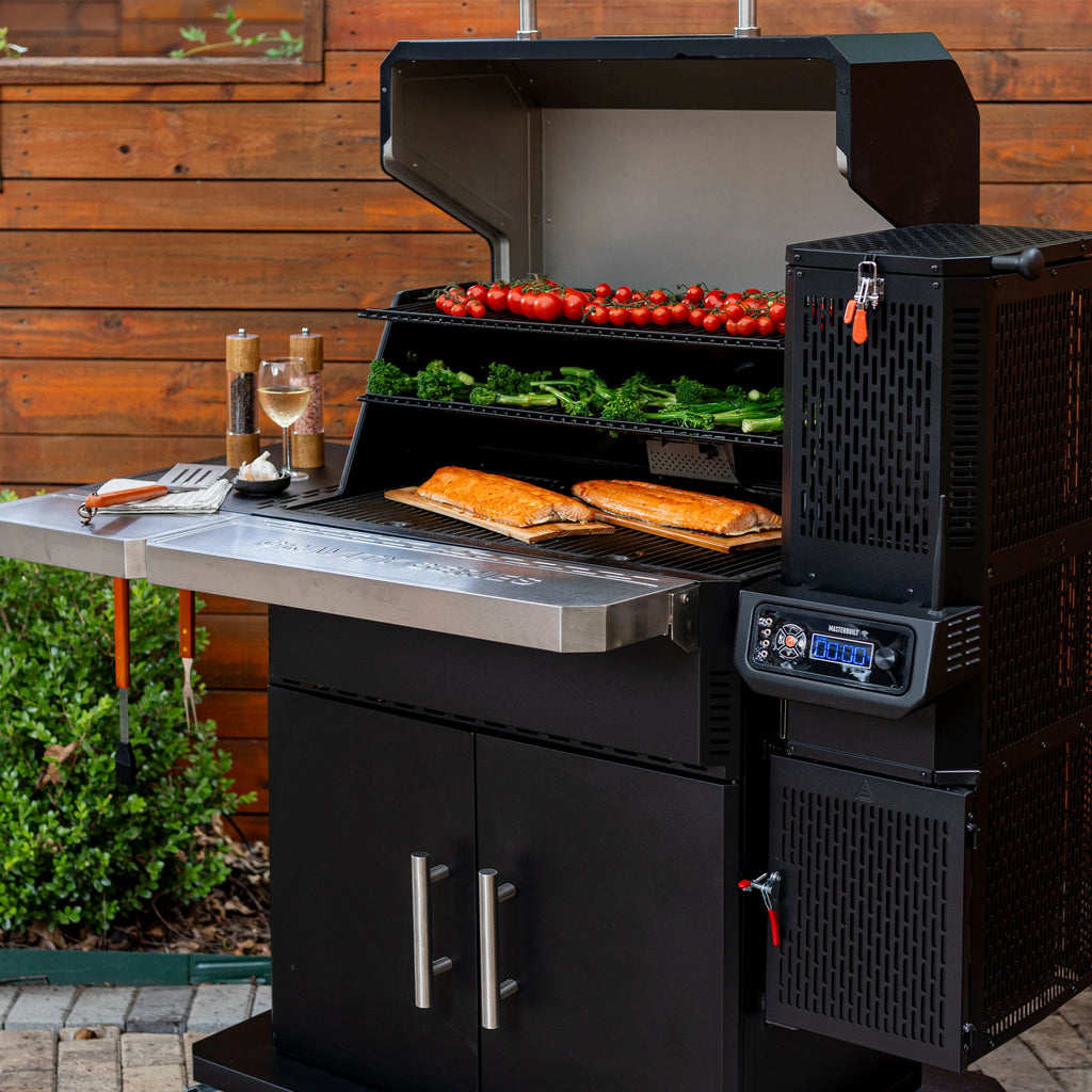 An open Gravity Series 1150 grill with the front shelf raised up. 2 salmon filets on wood planks cook on the main grate while broccolini and tomatoes on the vined cook on the warming rack. Utensils, salt and pepper grinders, and a glass of wine sit on the left side shelf.