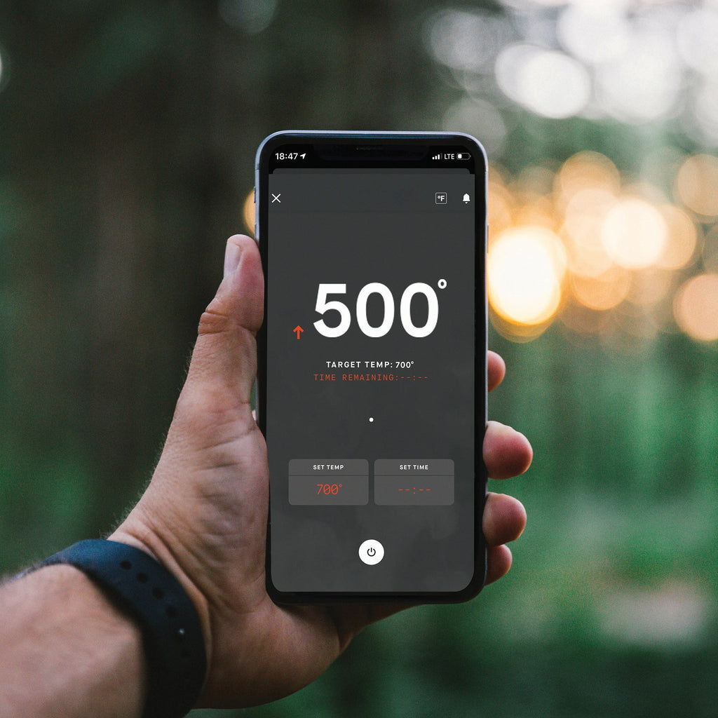 A man holds up a smartphone with the Masterbuilt app open. The app shows the current smoker temperature as 500 degrees with a target temperature of 700 degrees.