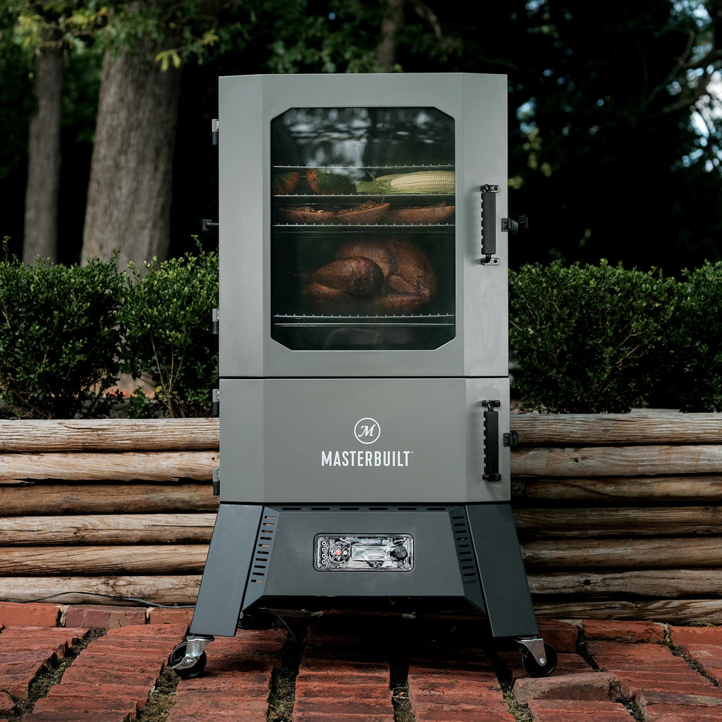 A full digital charcoal smoker sitting on a brick patio in front of a raised landscape bed made of small logs and filled with bushes.