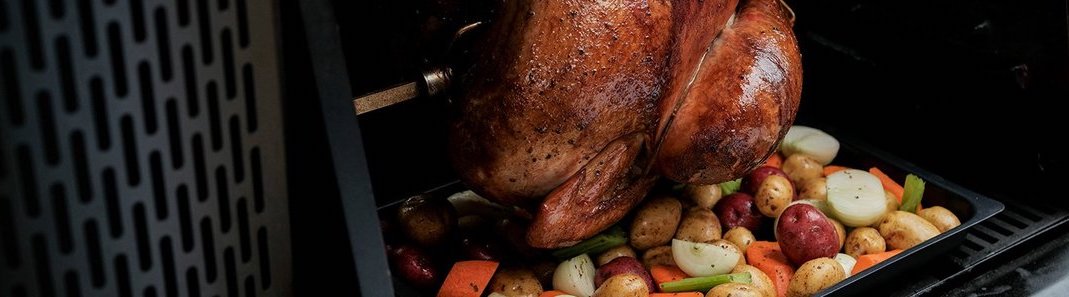 A turkey roasts on a spit over a tray of vegetables in a Gravity Series grill