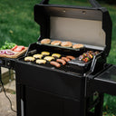 An open AutoIgnite Grill and Smoker. Cheeseburgers and sausages grill on the main cooking grate. 2 hamburger buns toast on the warming rack, and 2 skewers rest on the QuickSear hopper cast iron griddle plate. A wooden cutting board with lettuce leaves, sliced tomatoes, and grilled sliced onions is on the left side shelf. 