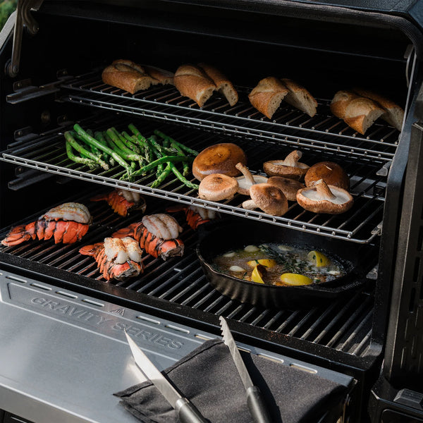 An open grill with 3 levels of food. Lobster tails and a cast iron skillet are on the bottom main cooking grate. Asparagus spears and whole mushrooms roast on the second tier. 4 buns open split-side down toast on the upper rack. A set of tongs and a towel rest on the front shelf.