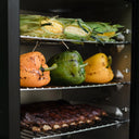 Closeup of 3 racks of smoked food: husk-on corn on the cob, whole bell peppers, and a rack of ribs.