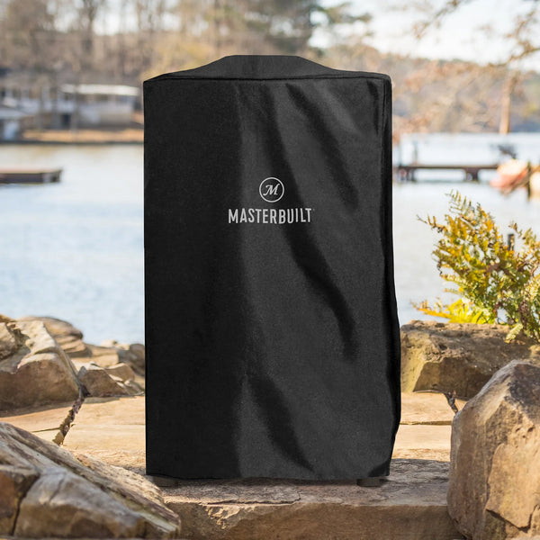 Black Masterbuilt grill cover over a vertical smoker sitting on a stone surface with a lake and trees in the background