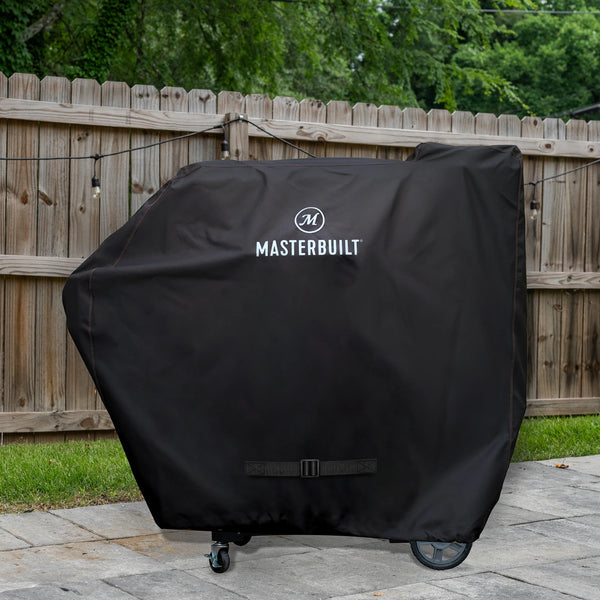 Black Masterbuilt grill cover on a patio with a wooden fence and greenery in the background