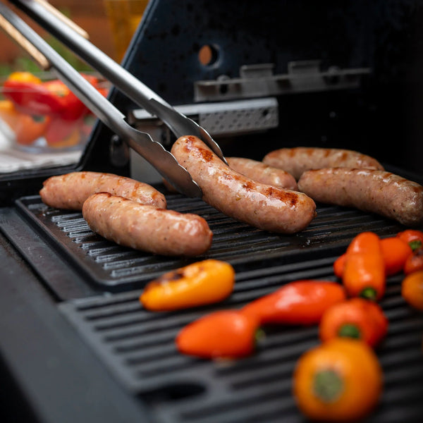 Someone using long metal tongs turns sausages searing on the ribbed side of a Flat-Top Griddle Accessory.