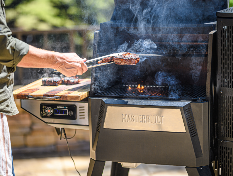A man moves seared steaks from the grill of a Gravity Series 1050 to a cutting board placed securely on the grill's side shelf