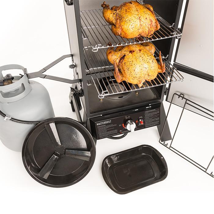 2 turkeys sit one per rack inside on open Masterbuilt Pro Series Dual Fuel Smoker. A small propane tank is mounted in a bracket to the left of smoker. Water and wood chip trays are displayed in front of the smoker.