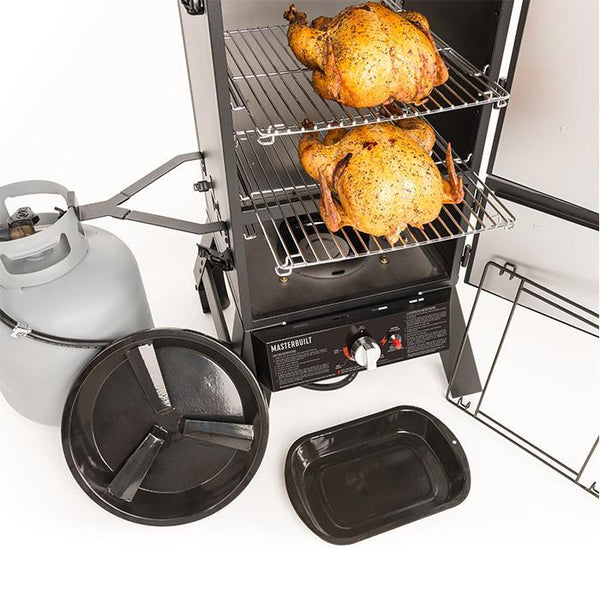 2 turkeys sit one per rack inside on open Masterbuilt Pro Series Dual Fuel Smoker. A small propane tank is mounted in a bracket to the left of smoker. Water and wood chip trays are displayed in front of the smoker.